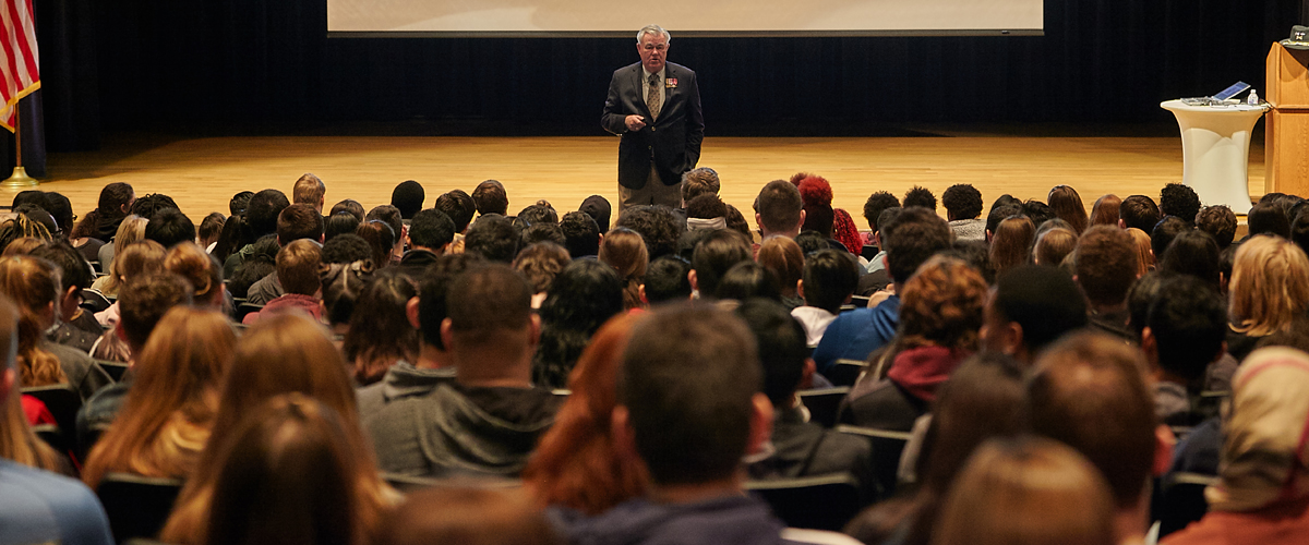 A crowd of students listen as a man speaks to them from a stage