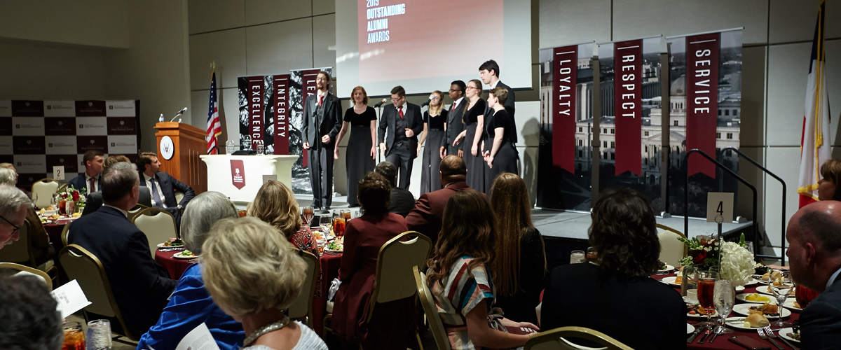 A choir group sings while on stage and a crowd is listening while sitting at tables