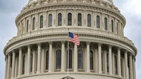 A close-up view of the United States Capitol dome, featuring its white columns and arched windows, with an American flag waving in the foreground against an overcast sky